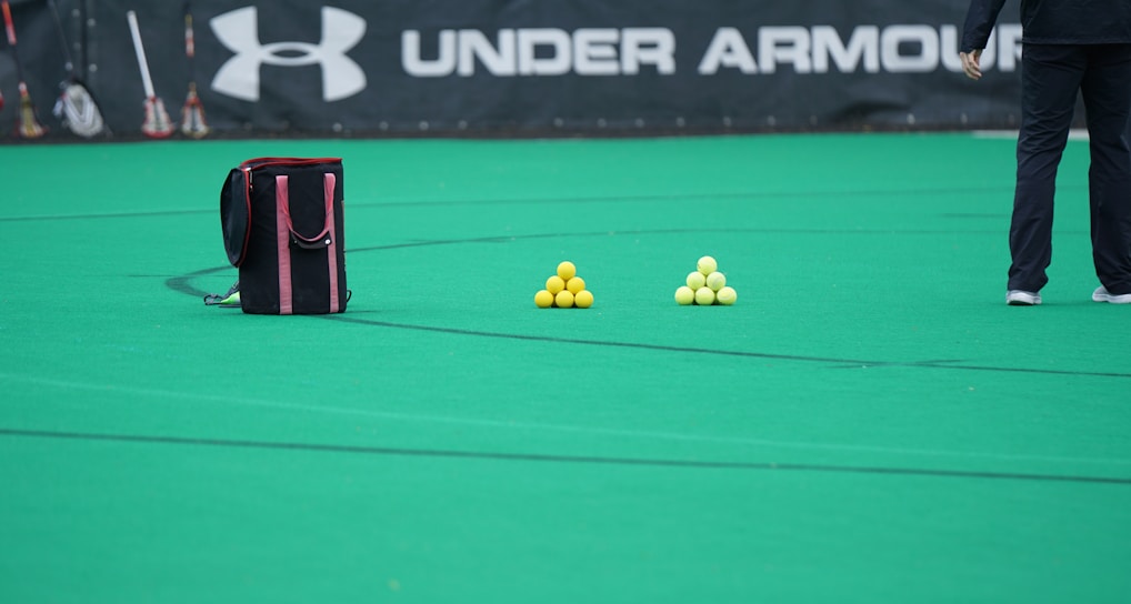 A sports field features a black and pink sports bag, two pyramids of yellow balls, and a portion of a person in dark athletic gear. The background displays a banner with the Under Armour logo.
