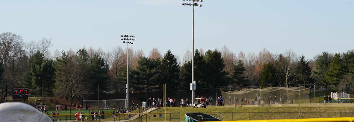 A sports field with a group of people engaging in athletic activities in the background. There is sports equipment like a soccer goal and a scoreboard visible. A chain-link fence runs across the foreground, and a statue or sculpture resembling an eagle is also seen. Trees and tall floodlights are positioned behind the field.