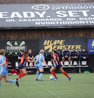 Players engage in a competitive soccer game on a field while others are seated on the bench watching attentively. The action is taking place in front of a wooden structure displaying multiple advertisements, including orthodontics, lacrosse, and field hockey.