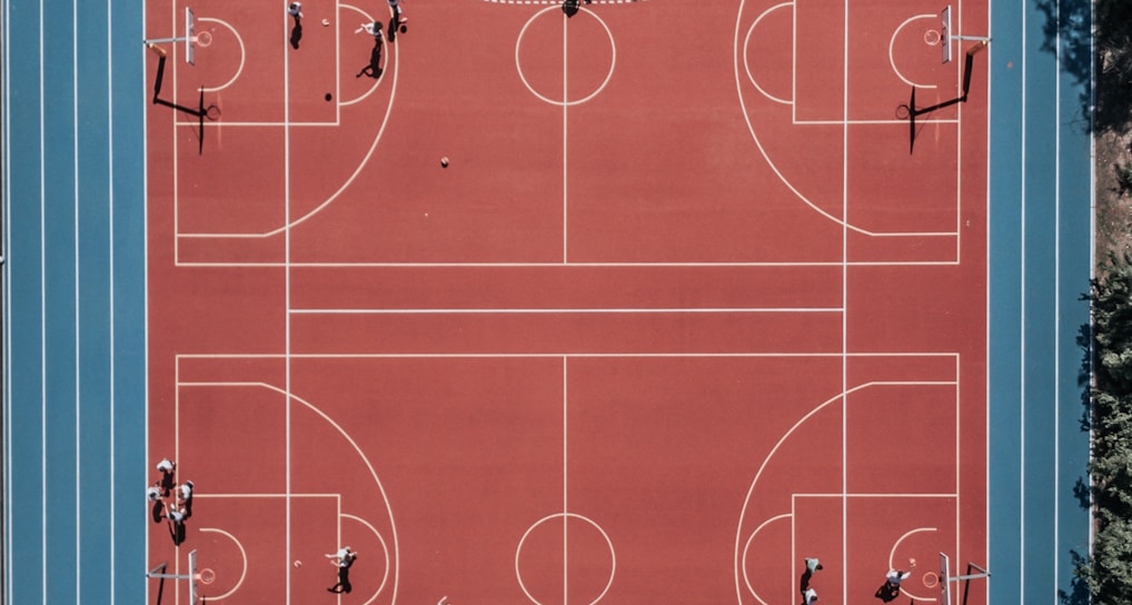 An aerial view of a large, vibrant sports complex featuring a red basketball court with multiple hoops and a surrounding blue running track. Several people are scattered across the area, engaged in various activities.