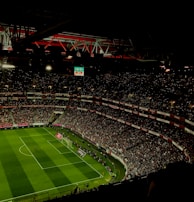 A large, illuminated stadium filled with a vast audience. The field is marked for soccer, with players actively participating in a match. The crowd is engaged, with numerous lights visible in the stands, possibly from phones. The stadium's roof structure is visible with red beams, and an electronic scoreboard shows a score of 0-0.