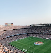 An expansive soccer stadium filled with thousands of spectators. The green field in the center features players and markings, surrounded by vibrant stands and advertising boards on the upper sides of the stadium. The audience is actively watching the game.