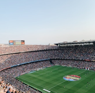 An expansive soccer stadium filled with thousands of spectators. The green field in the center features players and markings, surrounded by vibrant stands and advertising boards on the upper sides of the stadium. The audience is actively watching the game.