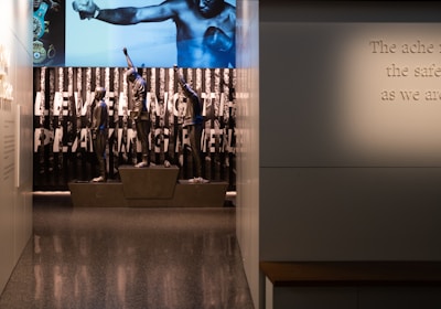 A dimly lit display features statues of athletes standing on a podium. They are positioned in front of a backdrop with the words 'LEVELING THE PLAYING FIELD' in bold letters. On the wall to the right, a softly lit quote is partially visible. The overall ambiance suggests a museum or exhibition setting.