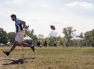 A person is engaged in a sports activity, running on a grassy field. The person is wearing a sports jersey and shorts. They are positioned in mid-air as they run, and a football is visible in front of them. In the background, there are several people along with tents and trees, indicating an outdoor sports event.