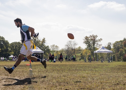 A person is engaged in a sports activity, running on a grassy field. The person is wearing a sports jersey and shorts. They are positioned in mid-air as they run, and a football is visible in front of them. In the background, there are several people along with tents and trees, indicating an outdoor sports event.
