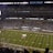 A packed football stadium with thousands of fans in attendance, centered around a brightly lit field. The field displays football markings and logos, indicating it's a professional game setup. An electronic scoreboard is visible, and the audience is engaged with the game.