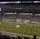A packed football stadium with thousands of fans in attendance, centered around a brightly lit field. The field displays football markings and logos, indicating it's a professional game setup. An electronic scoreboard is visible, and the audience is engaged with the game.