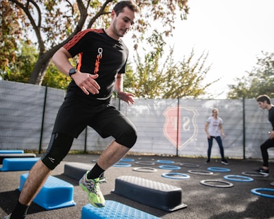 A man is actively participating in an outdoor fitness session, jumping over blue platforms with agility. Two other people in the background are also engaging in exercise, with rings scattered on the ground. The area is fenced and surrounded by trees, suggesting a park or recreational setting.