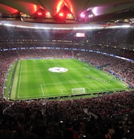 A large, packed stadium with bright lights illuminates a green soccer field. The scene is vibrant with a massive audience surrounding the pitch, visibly excited and engaged. The center of the field displays a prominent circular logo, suggesting a major sporting event. Players and staff can be seen on the field, likely preparing for a match.