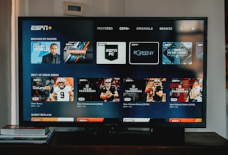 A television screen displaying the ESPN+ streaming interface with various sports-related shows and categories. The screen includes sections such as 'Browse by Shows' and 'Best of Drew Brees' with thumbnails of sports programs featuring athletes in action and analysts.