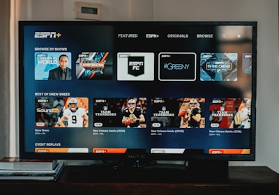 A television screen displaying the ESPN+ streaming interface with various sports-related shows and categories. The screen includes sections such as 'Browse by Shows' and 'Best of Drew Brees' with thumbnails of sports programs featuring athletes in action and analysts.
