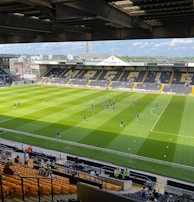 A sports stadium with a well-maintained grass field is depicted. Players are spread across the field, engaging in warm-up activities. The stands are partially filled with spectators, and the stadium is surrounded by urban buildings under a partly cloudy sky.