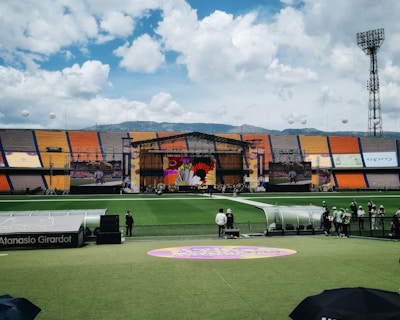A sports stadium with a large stage set up on the field, hosting an outdoor event. Spectators wearing colorful hats sit in the foreground. The seating areas display vibrant colors, and a clear blue sky with scattered clouds is visible. Large screens display graphics related to the event.
