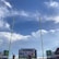 The image captures a view inside a large sports stadium, with bright yellow goalposts in the foreground reaching up towards a blue sky filled with fluffy white clouds. A crowd is present in the stands, partially visible underneath a large scoreboard displaying a sports game. Two spectators stand at the bottom center of the frame, with their hands raised, pointing upwards.