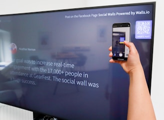 A person is holding a smartphone to take a picture of a large screen displaying a social media post. The content on the screen talks about increasing real-time engagement at a large event called GearFest. In the top corner, there's a QR code and a prompt to create a post.