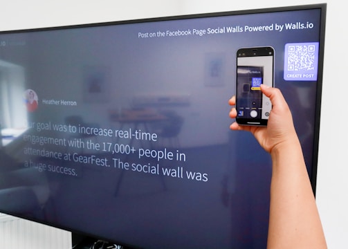 A person is holding a smartphone to take a picture of a large screen displaying a social media post. The content on the screen talks about increasing real-time engagement at a large event called GearFest. In the top corner, there's a QR code and a prompt to create a post.