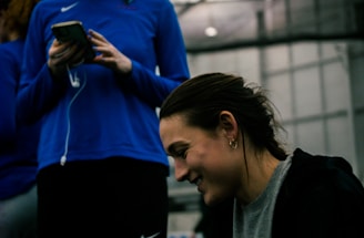 Two people are indoors in what appears to be a sports facility with a high ceiling and metal framework. One person is looking down at their phone with earphones in, wearing a blue long-sleeve athletic shirt. The other person is smiling and appears to be engaging with something or someone off-camera, dressed in darker athletic wear. The background is slightly blurred but shows additional people and possibly sports equipment.