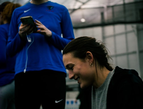 Two people are indoors in what appears to be a sports facility with a high ceiling and metal framework. One person is looking down at their phone with earphones in, wearing a blue long-sleeve athletic shirt. The other person is smiling and appears to be engaging with something or someone off-camera, dressed in darker athletic wear. The background is slightly blurred but shows additional people and possibly sports equipment.