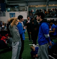 People engage in conversation and activities in an indoor sports facility. A woman in gray athletic wear stretches her arms above her head while talking to a man in a blue jacket. Other people are seen interacting in the background, some are seated on the ground, and the atmosphere is lively with many individuals gathered.