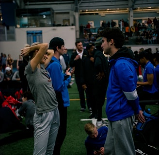 People engage in conversation and activities in an indoor sports facility. A woman in gray athletic wear stretches her arms above her head while talking to a man in a blue jacket. Other people are seen interacting in the background, some are seated on the ground, and the atmosphere is lively with many individuals gathered.