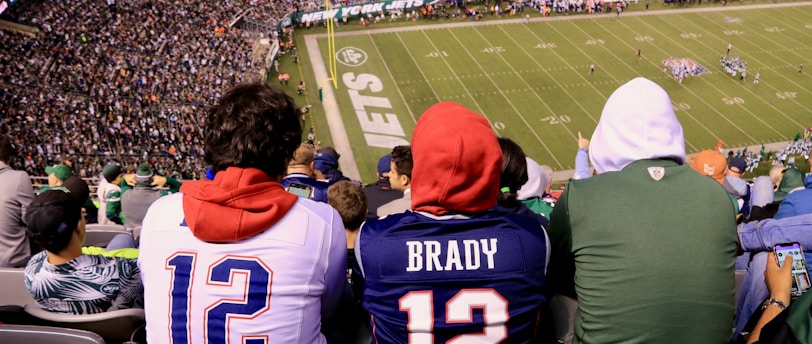 A crowded football stadium with fans sitting in the stands, wearing jerseys and hoodies. The field is visible in the background with teams lined up for a play. The audience is engaged, capturing the excitement of the game.